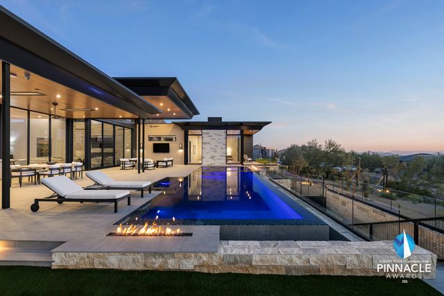The 2026 Pinnacle Award-winning pool by Morris Pools at dusk: a blue-lit vanishing-edge pool beside a modern stone-clad home, with a linear fire feature on a stacked-stone ledge