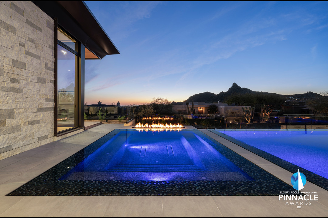 A blue-lit four-sided vanishing-edge spa with a line of fire behind it at dusk, desert peaks beyond