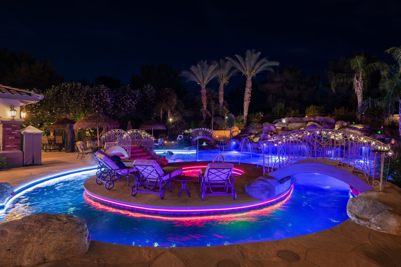 The 2026 PHTA Gold-winning freeform pool by Morris Pools at night: a lounge island ringed by a glowing lazy river, twin wrought-iron arched bridges, boulders and palms
