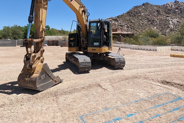 A CAT excavator on the graded lot, with the pool layout painted in blue and a rocky mountain behind