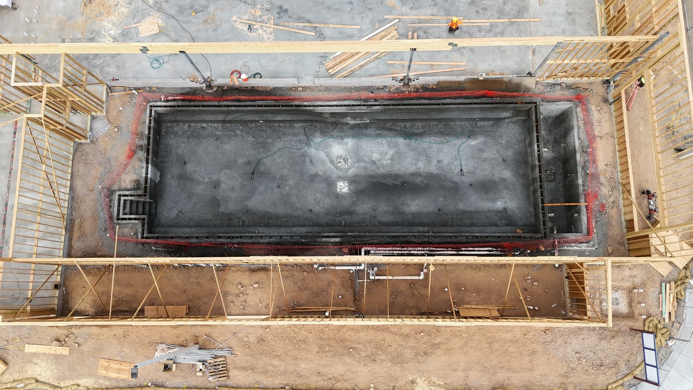Aerial view of a long rectangular pool shell with its perimeter gutter, framed by the walls of a building under construction