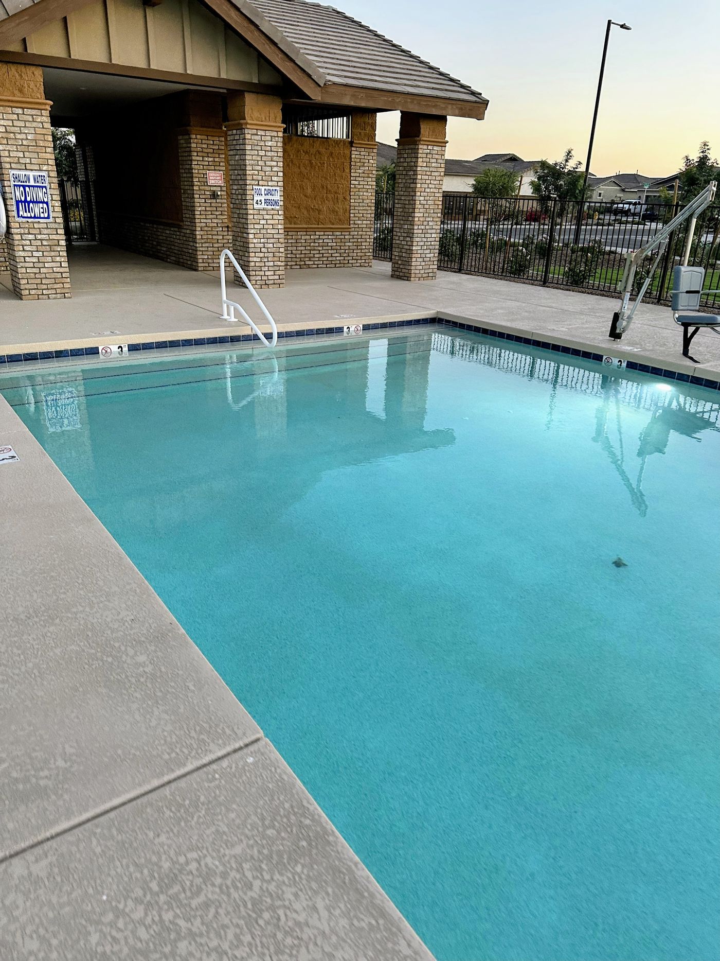 Community pool at dusk beside a brick pool building, with the tile line and accessibility lift in view