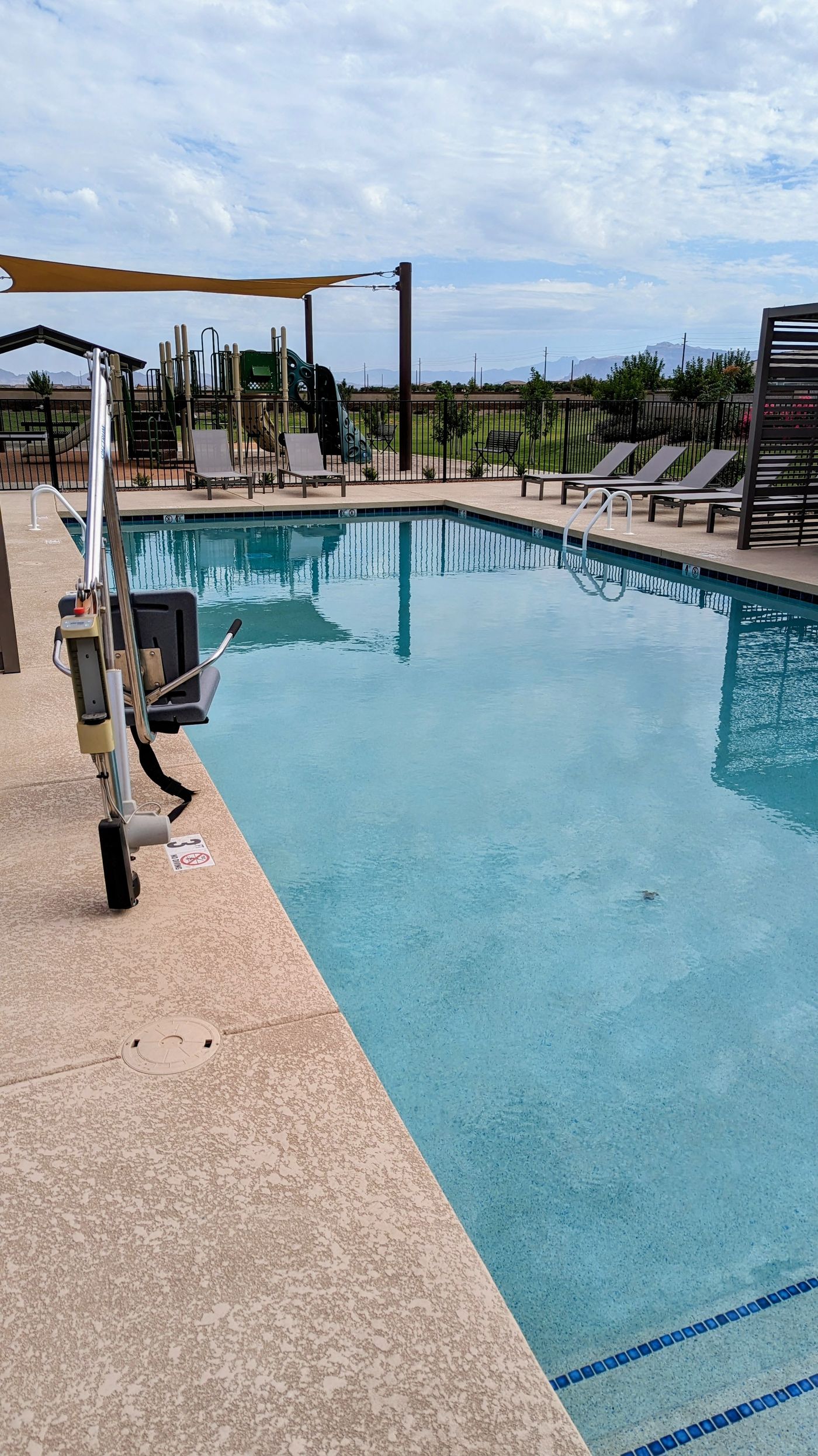 Community pool with an accessibility lift at the edge and a playground beyond the fence