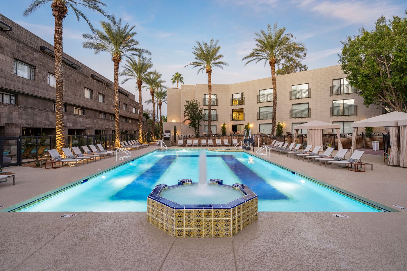 Resort pool at dusk with a raised octagonal fountain clad in patterned tile in the foreground, a deep-blue tile band inlaid in the pool floor, palms, loungers and white cabanas