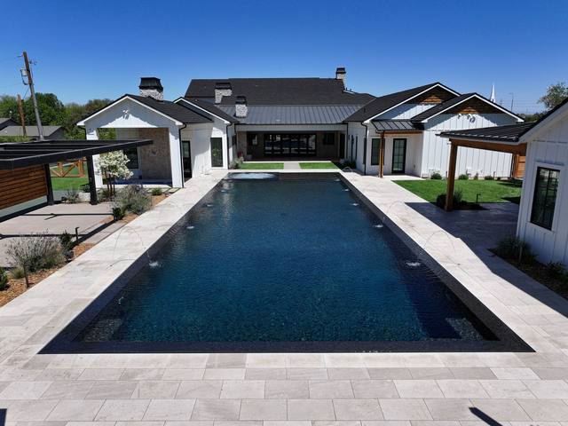 Long rectangular pool with black interior finish, deck jets and light pavers at a modern farmhouse