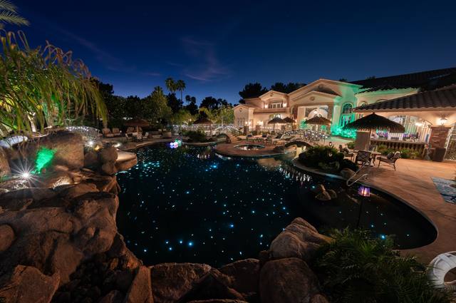 Lagoon-style freeform pool at night with fiber-optic starry floor lights, a rock grotto, bridges and palapas beside a Mediterranean estate
