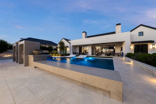 Wide dusk view of a modern pool with a raised spa wall, sun-shelf loungers and a fire feature in front of a white modern farmhouse
