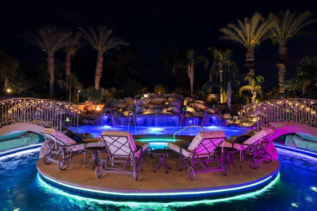 In-pool island with lounge chairs and LED-lit edges at night, reached by arched ironwork bridges, with a rock waterfall and deck jets under palms. 2026 PHTA International Awards of Excellence — Gold