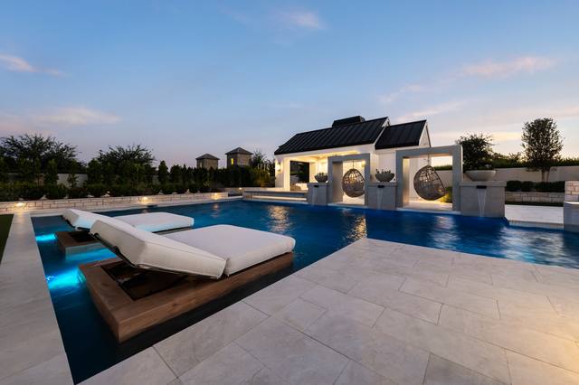 Modern pool at dusk with loungers on an in-water sun shelf, travertine-style pavers and a pavilion with hanging egg chairs